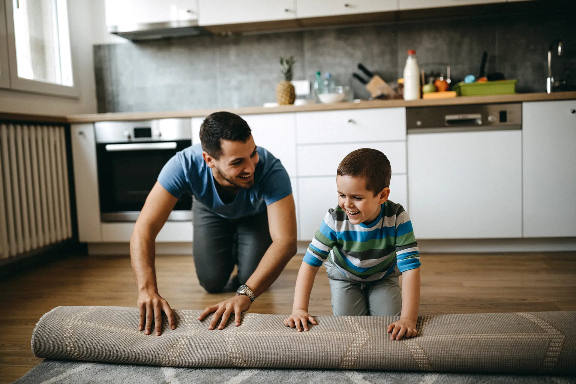 Man and his son having fun unrolling a new rug on their apartment floor.