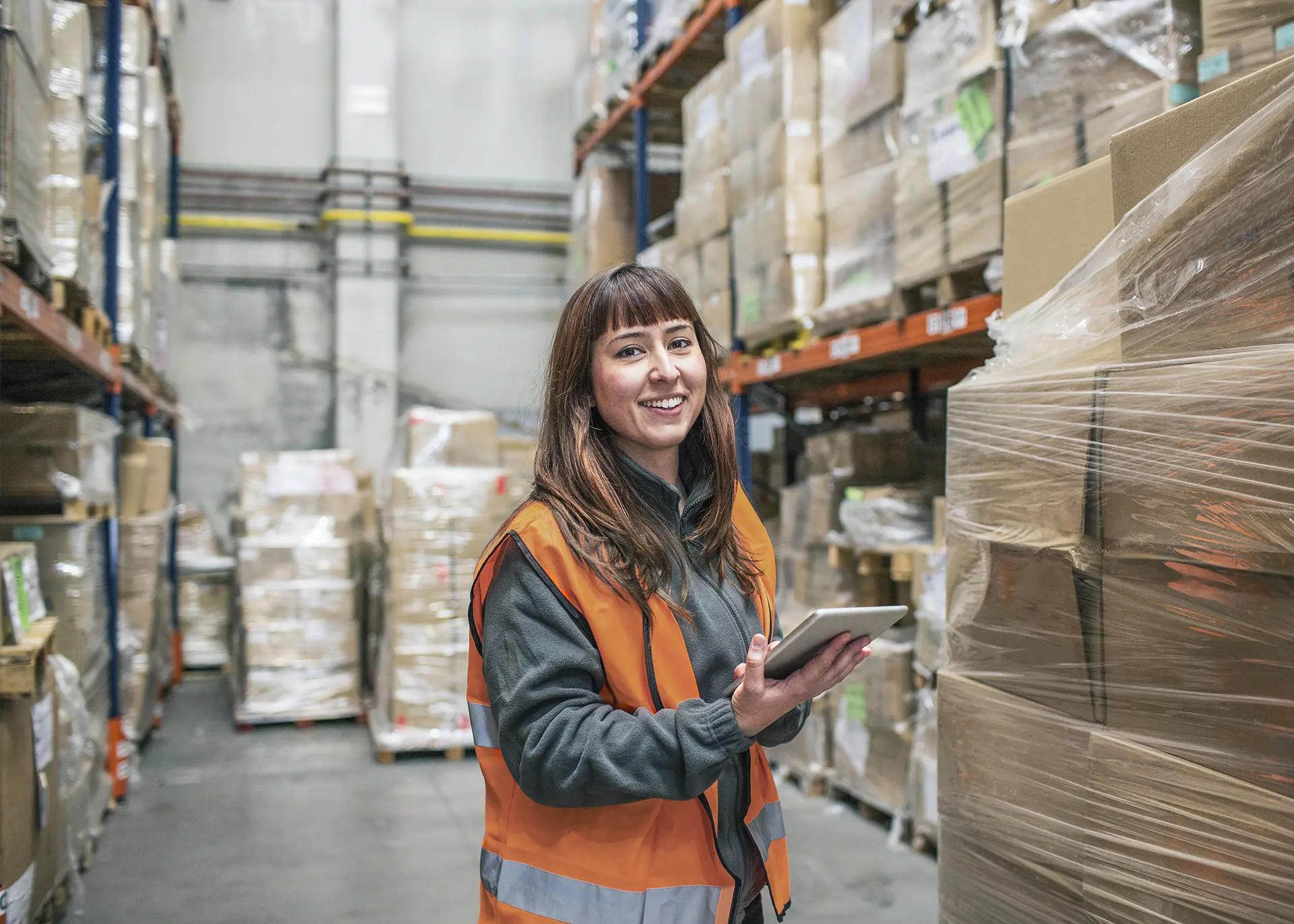 A woman in an orange vest smiling and using a tablet to track plastic wrapped boxes going out for shipment